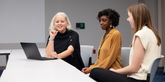 1 woman sits at a laptop while 2 women smile and discuss 