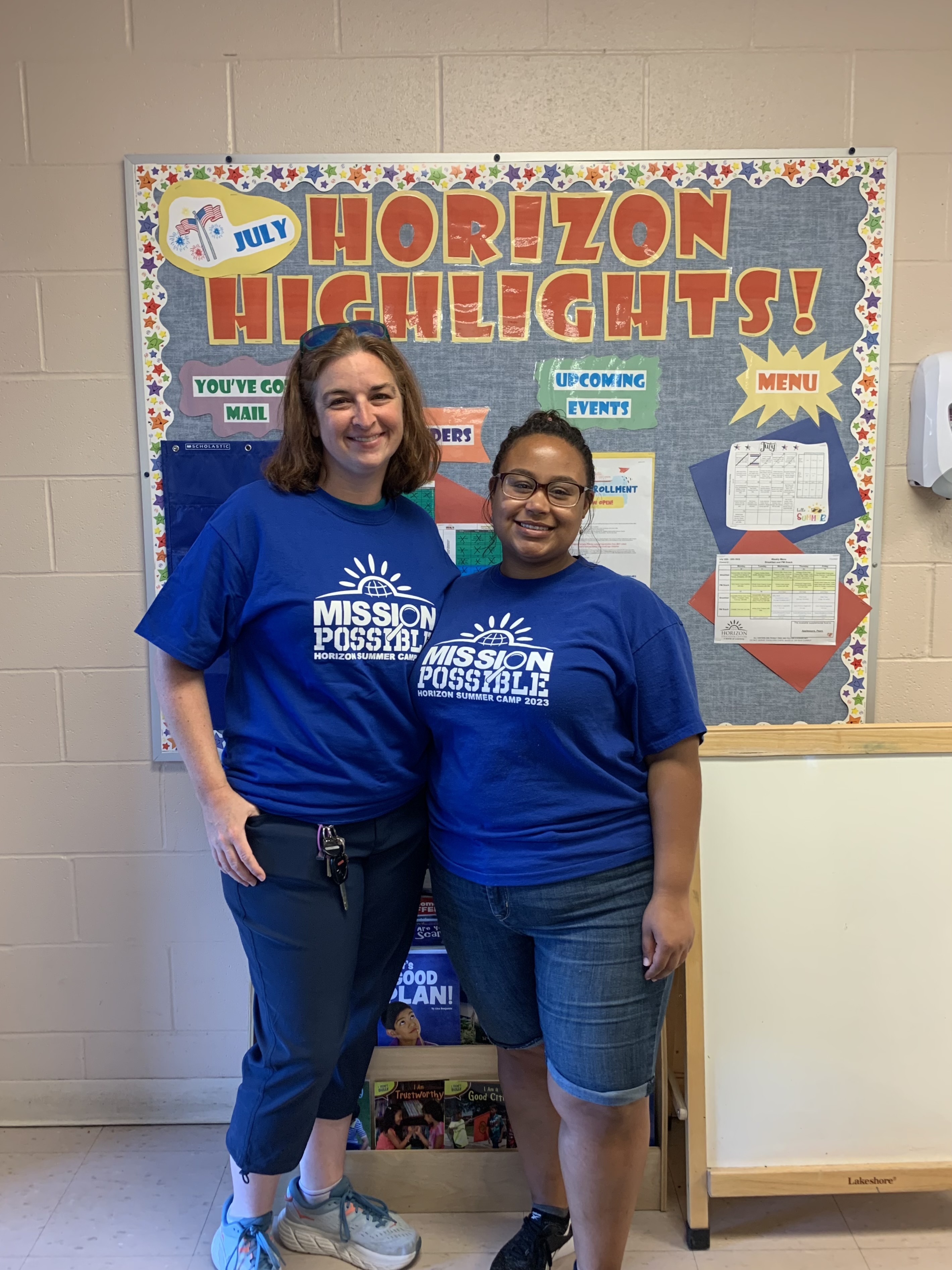 two women standing next to a bulletin board at an early childcare center